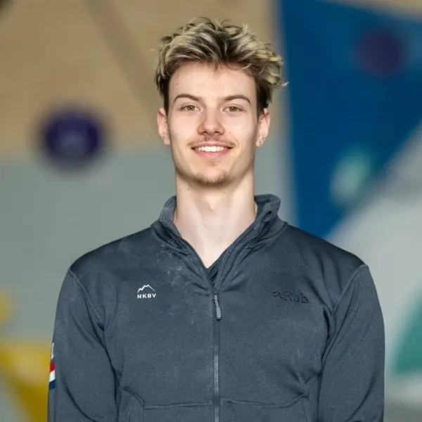 Een jonge man met kort, stekelig blond haar glimlacht naar de camera; hij draagt een grijs jack met ritssluiting en een klein logo op de borst. De achtergrond is wazig en bestaat uit blauwe, beige en 